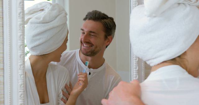 Couple smiling and enjoying time together in bathroom. Both appear in white robes, one with a towel wrapped around head and holding a toothbrush. This image can be used for promoting self-care, personal hygiene products, or relationship-building themes.