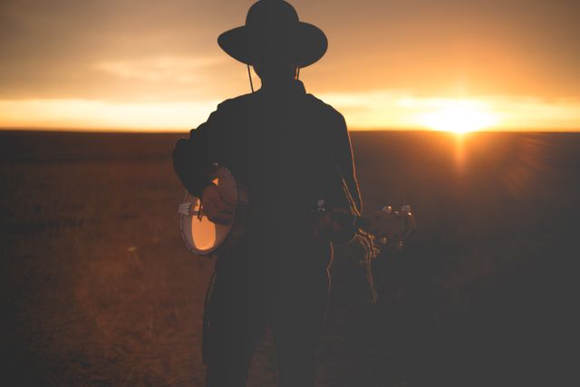silhouette of a man wearing cowboy playing a guitar in grass field against sunset sky. Country music concept