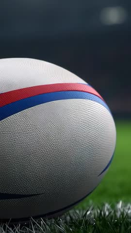 Vertical rugby opener showing textured ball resting on grass under stadium floodlights