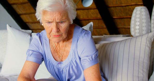 Elderly woman with short grey hair sitting on a couch looking worried and thoughtful. She is wearing a light purple blouse. The scene suggests a moment of reflection, concern, or decision-making. Useful for topics on elderly care, mental health, loneliness, and retirement.