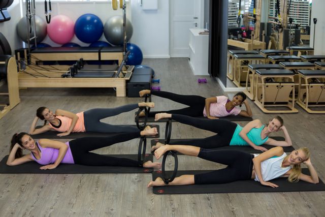 Five women are lying on their sides on exercise mats in a gym, using Pilates rings for their workout. They appear focused and coordinated. The background includes exercise balls, Pilates equipment, and mirrors. This image can be used for promoting group fitness classes, pilates training, health and wellness programs, or gym memberships.