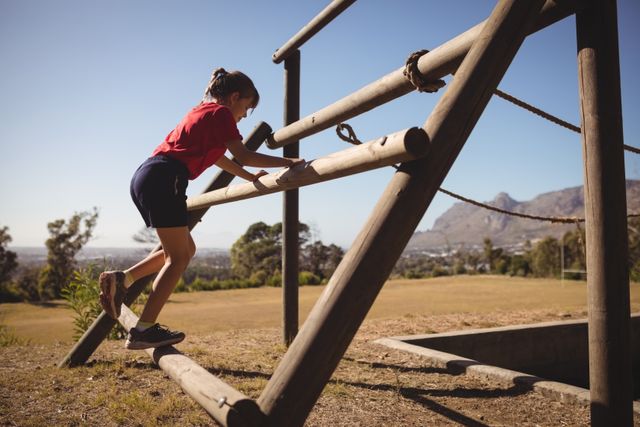 Young girl climbing wooden obstacle course in outdoor boot camp. Ideal for illustrating children's fitness, outdoor activities, summer camps, physical education, and healthy lifestyle. Perfect for use in educational materials, fitness blogs, and promotional content for youth sports programs.