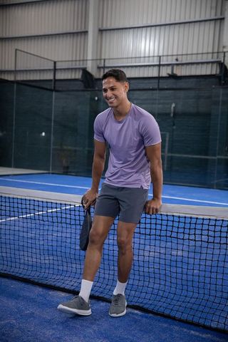 Asian man leaning on net at indoor padel court holding paddle smiling in athletic wear