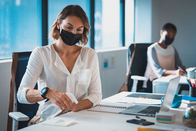 Two diverse businesswomen wearing face masks sitting in office, one disinfecting her desk. independent creative business at a modern office during coronavirus covid 19 pandemic.