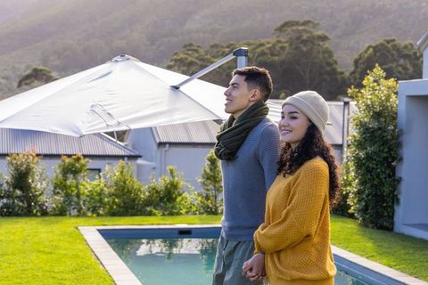 Asian couple holding hands by backyard pool, smiling, enjoying mountain view in sweaters