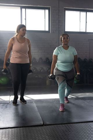 Diverse women working out lifting dumbbells doing lunges in industrial gym studio