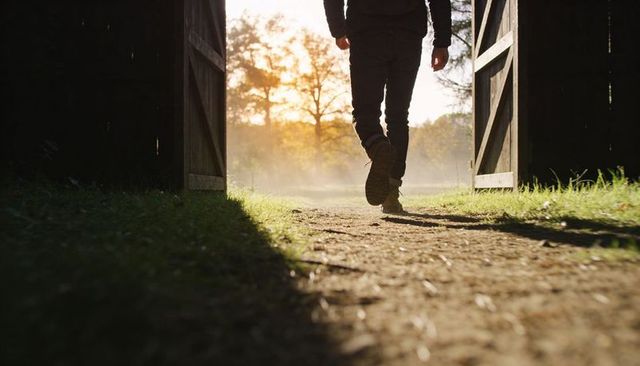 Backlit man walking through barn doorway into golden hour mist with hiking boots