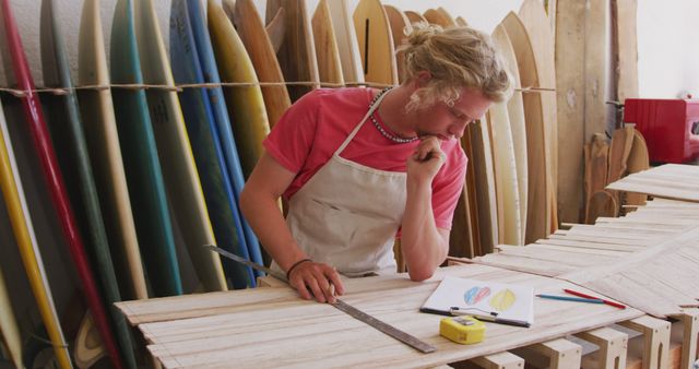 Young man engaged in shaping a surfboard in professional surfboard workshop. Image shows close-up of craftsman measuring with ruler and notes. Ideal for articles on surfboard making, craftsmanship, detailed processes, or focusing on artisans at work.