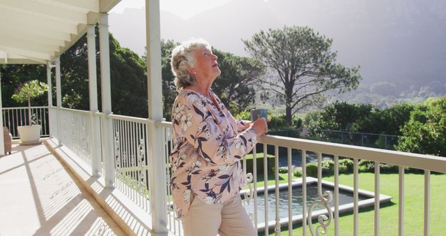The image shows an elderly woman standing on a balcony, enjoying her drink while overlooking a pool and lush garden scenery. This could be used in promotions for retirement homes, lifestyle blogs about aging gracefully, or articles about enjoying nature and relaxation in one's later years.
