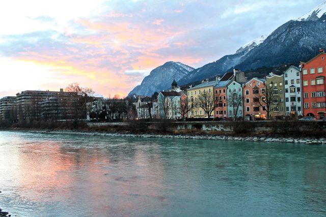 Colorful buildings by the river with mountain background reflects beautiful lights during sundown. Ideal for illustrating the charm of European towns, travel guides, tourism brochures, architectural designs, and urban planning presentations.