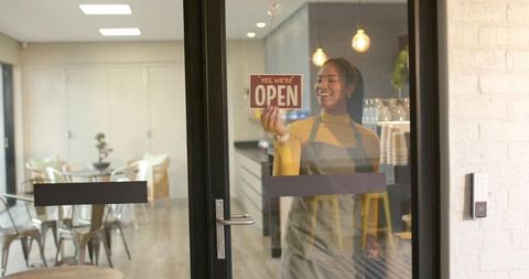 African american woman hanging open sign, smiling in modern cozy cafe interior