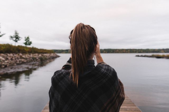 Woman with a ponytail standing on a dock by the lake, looking into the distance. The sky is overcast and she is wrapped in a blanket suggesting cool weather. This image is ideal for representing serenity, peace, contemplation, and the beauty of outdoor nature settings. It can be used in websites, advertisements, and educational materials related to nature, travel, mindfulness, and relaxation.