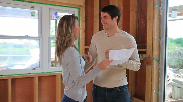 A couple stands in a partially renovated room, smiling at each other while holding construction plans. This moment reflects joy and teamwork as they embark on a new chapter together. The atmosphere is one of excitement about their home's potential. Ideal for real estate, home improvement themes or lifestyle visuals demonstrating partnership and new milestones.