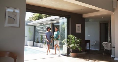 Walking couple carrying yoga mats through folding glass doors into sunlit poolside patio