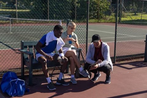 Multiethnic tennis players taking break and reviewing tablet courtside on sunny outdoor court
