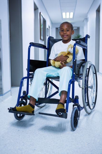 Portrait of boy sitting on wheelchair and holding teddy bear in corridor at hospital