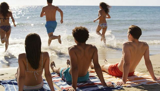 Friends relaxing on striped towels watching group running into sunlit surf