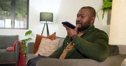 African American man speaking into smartphone while relaxing on sofa in cozy living room
