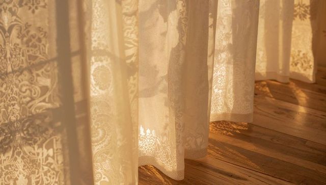 Sunlit cream lace curtains casting delicate floral shadows on warm wooden floorboards