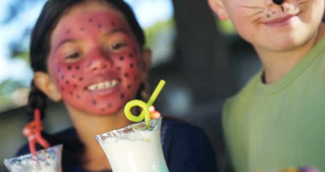 Two children with face paint enjoy a playful moment, with copy space. The girl with a ladybug design and the boy with a cat whiskers design are holding glasses with a straw, suggesting a festive or party atmosphere.