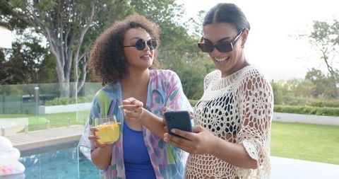 Two african american women laughing by poolside, checking phone and enjoying drinks