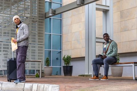 Diverse young men waiting with suitcase and phone on urban bench near modern architecture