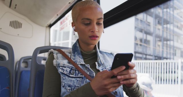 A young woman with short blonde hair and earphones is using a smartphone while traveling on a city bus. She is wearing a denim jacket over a green top and appears focused on her device. This image is ideal for content relating to public transportation, commuting, modern technology, urban living, and connectivity in everyday life.