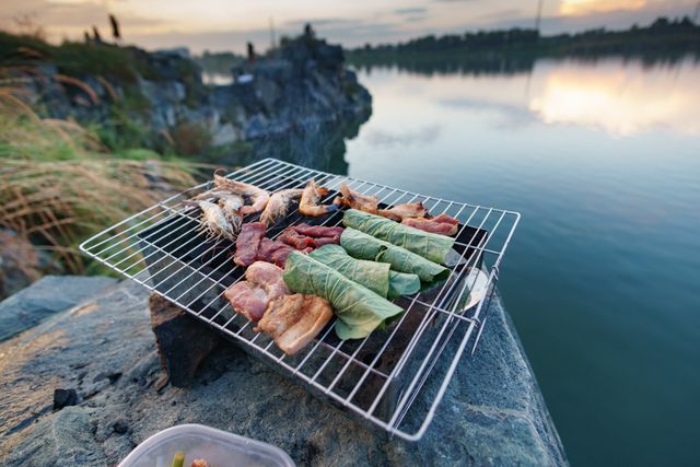 Barbecue set near lake at sunset with various meats and vegetables on grill. Perfect for use in articles, blogs, or marketing materials related to outdoor cooking, leisure activities, camping, and summer adventures. Ideal for promoting products and services related to travel, food, and lifestyle.
