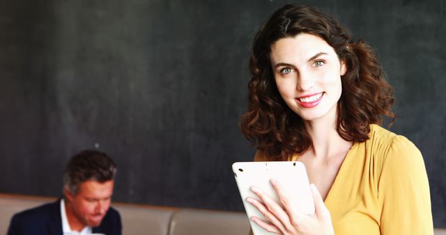 Confident woman holding tablet and smiling with a man working in the background. Great for depicting modern work environments, female leadership, professional settings, business concepts, and technology use. Ideal for articles and promotions focused on career growth, business successes, and teamwork.