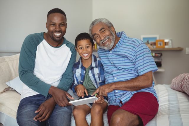This image shows a joyful multi-generation African American family bonding over a digital tablet while sitting on a bed in a bedroom. The grandfather, father, and son are all smiling and enjoying their time together, highlighting themes of family, technology, and leisure. This image can be used for advertisements, articles, or blog posts related to family life, technology use in households, or intergenerational relationships.