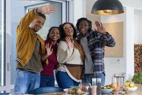 Diverse friends smiling and waving around dining table during casual home gathering