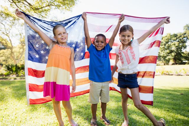 Three happy children are holding a USA flag in a park, smiling and enjoying a sunny day. This image can be used for themes related to patriotism, diversity, childhood joy, and outdoor activities. It is ideal for advertisements, educational materials, and social media posts celebrating national holidays or promoting unity and friendship.