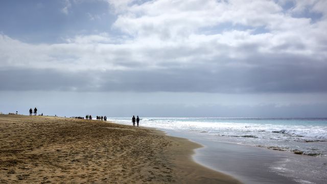 Several people walking along a sandy beach under a cloudy sky. Ideal for themes related to leisure, beach walks, seaside activities, outdoor relaxation, nature exploration, travel, and vacation.