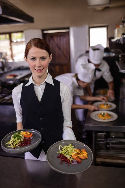 Female waitress holding plates with gourmet food in a professional restaurant kitchen. Chefs in the background are preparing dishes, showcasing teamwork and culinary skills. Ideal for use in articles or advertisements related to the hospitality industry, restaurant services, culinary arts, and fine dining experiences.