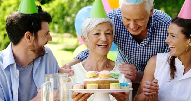 A diverse group of adults is celebrating outdoors, with a senior Caucasian man and woman enjoying a birthday cake with a younger Caucasian couple, all wearing party hats. Smiles and a festive atmosphere convey a sense of joy and family togetherness during a special occasion.