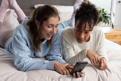 Two friends sharing smartphone on bed laughing and browsing social media together