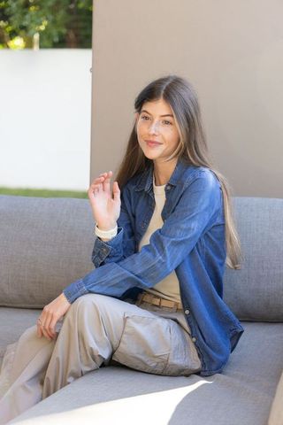 Young woman waving on sunlit patio sofa, casual relaxed outdoor lifestyle portrait