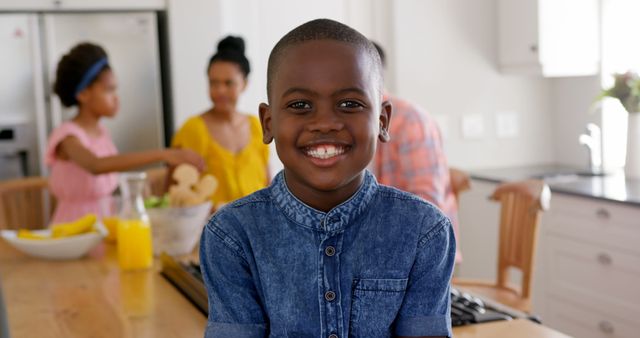 Young boy smiling in a well-lit kitchen with his family in the background. Suitable for family lifestyle, childhood happiness, and home life themes. Ideal for advertisements, magazine articles, and social media posts focusing on happy family moments and child development.