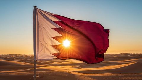 Qatar flag waving over rippled desert dunes at sunset with dramatic sunburst backlight