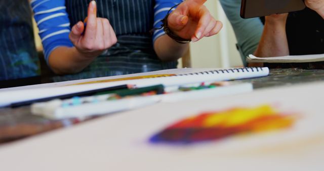 Artists in casual clothing work together on a colorful abstract painting in an art class. They are pointing and discussing techniques while looking at a tablet and sketchbook on a table filled with other art supplies. This image can be used to depict creativity, collaboration, art education, and teamwork in artistic projects.