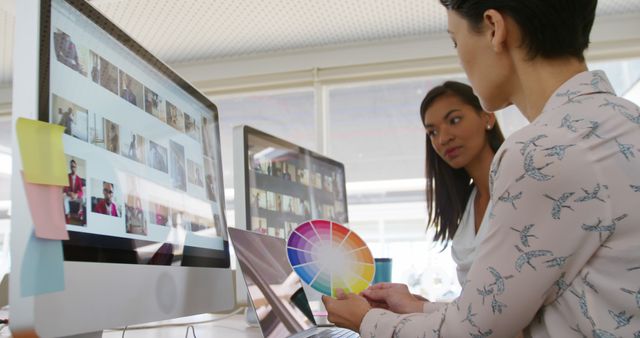 Two women are actively engaged in a design discussion, examining a color wheel and images on computer screens in a modern office. This setting highlights themes of creativity, teamwork, and project planning, making it ideal for business, technology, or creative industry promotional materials. The image can be used in articles or advertisements focused on graphic design, digital media, and professional collaboration.