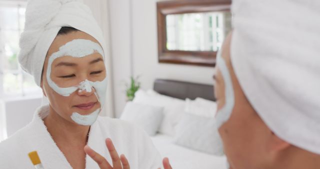 Asian woman in white bathrobe and towel wrap applying facial mask in front of mirror. Can be used for promotions related to skincare products, home spa routines, beauty and self-care tips, or wellness articles.