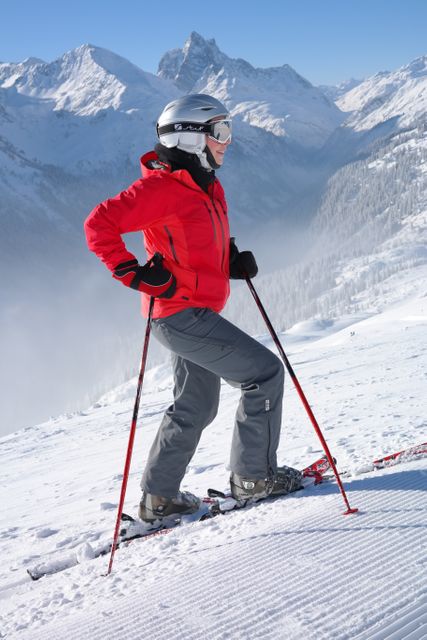 Person standing on skis with ski poles, wearing red jacket and helmet, high in mountains surrounded by snow-covered peaks. Ideal for use in advertisements for winter sports equipment, travel brochures, outdoor adventure magazines, and promotional materials for ski resorts. Showcases the thrill and beauty of winter sports with a picturesque mountainous backdrop, offering inspiration for outdoor enthusiasts.