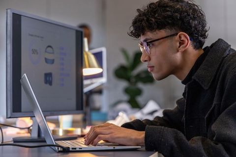 Young professional analyzing data on laptop with dual monitors in modern coworking office