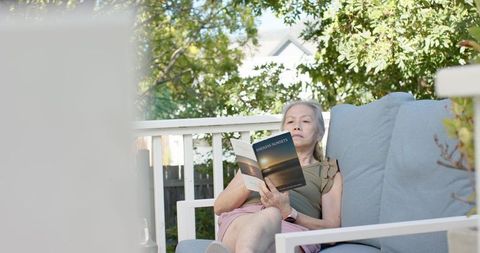 Senior asian woman reclining on porch sofa reading novel endless sunsets during afternoon
