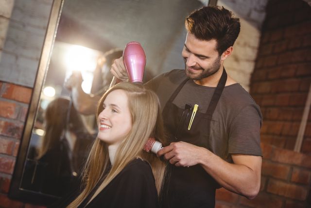 Woman sitting in a hair salon while a hairstylist dries her hair with a blow dryer. The hairstylist is wearing an apron and smiling, creating a friendly and professional atmosphere. This image is perfect for use in beauty and hair care advertisements, salon promotions, and articles about hair styling and grooming.