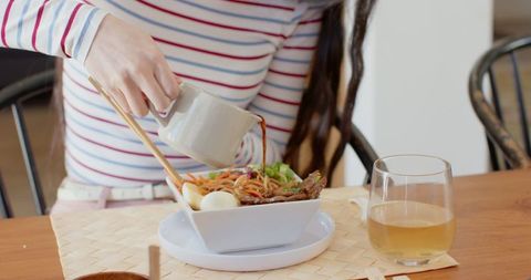 This image shows a woman pouring sauce onto a bowl of Asian noodles with chopsticks on a casual dining table. It includes a glass of tea, contributing to a warm and cozy atmosphere. Suitable for depicting culinary experiences, food blogs, restaurant promotions, and cooking tutorials emphasizing cozy and relaxed culinary settings.