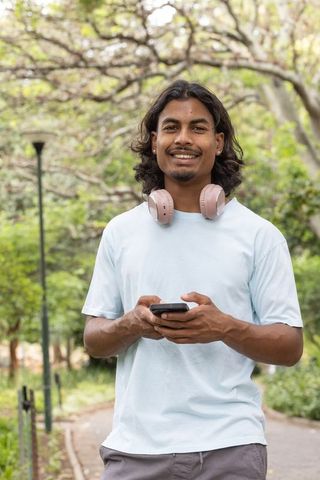 Smiling african american man standing in park holding smartphone wearing pink headphones