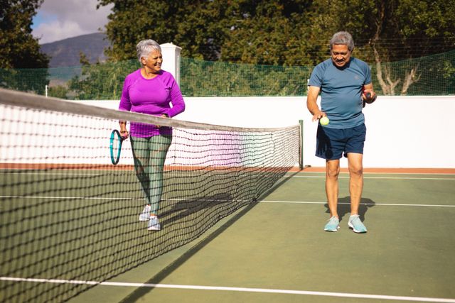 Biracial senior woman looking at senior man playing with tennis ball by net at court in summer. sport, competition, unaltered, love, togetherness, lifestyle, retirement and active lifestyle.