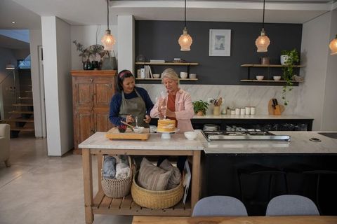 Mature friends baking together piping frosting on layered cake in modern marble kitchen